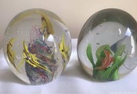Photo showing two aquatic-themed glass paperweights side by side, one with yellow and multicolored fish design, the other green aquatic plant design with bubbles.