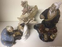 Overview of three ceramic Asian figurines: two elderly men playing a game, single seated man, and floral bird background piece.