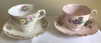 Two tea cups and saucers displayed side by side. One set white with multicolor flowers and gold trim, the other pink with purple flowers and gold trim.