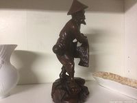Wood carving figurine of a man holding a basket wearing a conical hat, standing on a carved base.