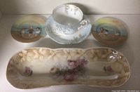 Top view of vintage dishware including two decorative plates with children and horse scenes, a floral teacup and saucer, and a rectangular floral platter.