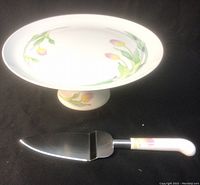 Pedestal cake plate on black background alongside matching cake lifter with floral handle.
