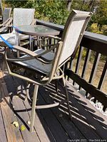 Two bar-height swivel chairs with metal frames and beige mesh fabric around a 28-inch round glass top bar-height table with metal frame and umbrella hole, on a wooden deck outdoors.