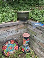 Ceramic bird bath base with colorful floral and geometric patterns, next to a large green glazed planter on stone steps outdoors.