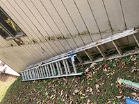 Two extension ladders (24 ft and 30 ft) resting next to a garage wall on the grass, tied together with a blue strap, with leaves around them.