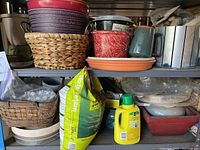 Shelves containing several different planters including woven basket, ceramic red pot, and purple plastic pot. Bag of fertilizer and a yellow bottle of plant food visible in front.