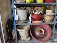 Multiple ceramic and plastic planters arranged on metal shelving in garage, including large beige planter, red planter, and various sized pots.