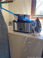 Roaster oven, mixing bowl and iron shown on top of a white cabinet with window background.