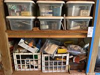 Furniture shelf with clear plastic bins and white crates filled with assorted hardware items such as screws, nails, electrical parts, and door knobs.