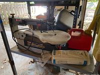 Side view showing Craftsman scroll saw with cord, red plastic fuel container behind it, and 3 rolls of clear plastic sheeting in packaging on shelf below.