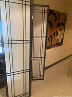 Full view of three-panel privacy screen leaning against wall showing black frames and white paper inserts with geometric black pattern.