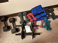 Photo showing assortment of variously sized handheld dumbbells in black metal and colored neoprene coatings, two dumbbell bars, and a blue ab roller.