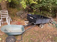 Wide view showing the yard cart with black container and plastic wheelbarrow in outdoor garden area with grass and fallen leaves.