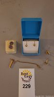 Assorted items on table including red gemstone studs in plastic case, blue jewelry box with floral earrings, two square rings, gold T-bar chain, single earring element and lot tag