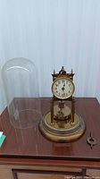 Full view of the antique anniversary clock showing brass base, clock face on two pillars, glass dome cover beside it and key lying on wooden surface.
