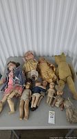 Lot overview showing 7 antique dolls along with a plush teddy bear and plush cat on grey table