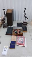 Full view of two bronze stork candlesticks, various books including Bibles and hymnals, and boxes of playing cards and game pieces arranged together on a table.