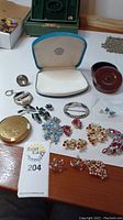 Photo showing assortment of costume jewelry including brooches and clip earrings with multicolored semi-precious stones along with vintage-style makeup compacts and a leather case.