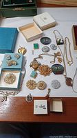 Overview of all costume jewelry items spread on a table with boxes, showing variety of pins, brooches, necklaces, and earrings in gold and silver tones with colored stones.