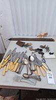 Photo showing a collection of yellow-handled knives, forks, spoons, metal letter stamp with wax seal, multiple keys and locks, and two leather barber straps on a table.