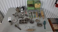 Overhead view showing the green metal toy stove and assortment of vintage metal kitchen accessories including kettle, coffee pot, cups, rolling pins, spatulas, ladle, muffin pan, cookie cutters, and a plastic scoop all laid out on a flat surface.
