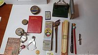 Full layout of various vintage office tools laid out on a flat surface showing tins, compass, letter openers, small book, measuring tool, and green desktop hole punch or stamp.