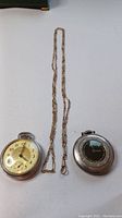 Two Westclox pocket watches and a linked metal chain laid flat on white surface.