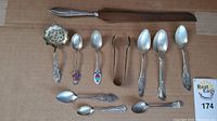 Full view of 11 sterling silver utensils laid out on brown background including spoons, tongs, and cutting utensil with stainless steel blade