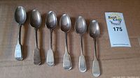 Seven spoons arranged in a row on cardboard with lot tag