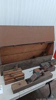 Photo showing large rusted metal toolbox open revealing wooden interior lining and four wooden hand planers on table.