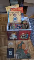 Top-down view of cardboard box containing various children's books with some titles visible at cover and additional books laid out in front on floor