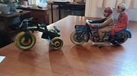 Green tin tricycle with radial color wheels next to blue tin motorcycle with two riders on wooden surface