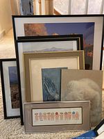 Seven framed art prints stacked leaning on each other on carpet, varying frame styles and sizes visible