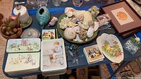 Overview of bathroom and sea themed decor on blue table including cactus, ceramic vase, seashells, framed artwork, collectible tins, and coasters.