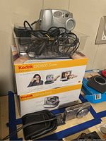 Kodak DX3600 Zoom digital camera, Kodak Camera Dock box, and accessories including cables and manual in plastic container seen on a surface.