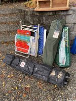 Photo showing four folded colorful folding chairs and two green canvas bags for canopy, placed outdoors on gravel with stone wall background.