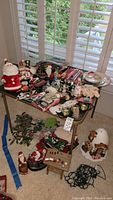 Wide view of table covered with assorted Christmas decorations including Santa Claus figures, candles, faux holly, snowmen, decorative plates, stockings, and other holiday items.