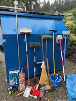 Photo of assorted push brooms, mops, buckets, whisk broom, scrub brushes, and small cleaning tools placed against a blue dumpster outdoors.