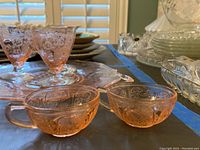 Photo showing two pink glass cups, a large pink serving platter with floral patterns, and parts of other glass items in the background.