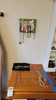 Full view showing several necklaces hanging on a decorative wall hanger above a wooden surface with more necklaces laid out.