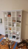 Full front view of white cubby shelf showing 24 square compartments and various small objects inside shelves and on floor in front of it.