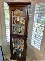 Full view of wood curio cabinet with two glass door sections, displaying plates and figurines inside the glass shelves. Cabinet has classic molding detail on top and warm brown finish.