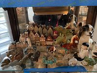 Figurines displayed on a shelf including Asian ceramic figures, ceramic house village set, and multiple animal figurines made of stone and ceramic.