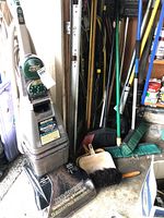 Hoover Steam Vac vacuum cleaner and multiple cleaning accessories including brooms, dust pan, hand brush, and clip-on spout shown together in a garage setting.