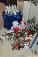 Wide image showing white nativity set figures arranged on a blue velvet cloth, Santa Claus and Mrs. Claus dolls, various stuffed animals including snowmen, reindeer, and teddy bears, several Christmas trees, and holiday pillows.