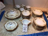 Overview of Noritake china set with stacks of plates, saucers, cups, and two covered serving dishes arranged on carpeted floor.