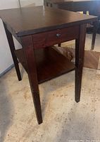 Front angle view of the wooden side table showing drawer and lower shelf, with visible scratches and scuffs on legs and surfaces.