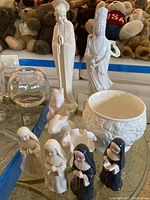 Photo showing statuettes including Madonna, Kuan Yin, nuns, bunnies, hobnail candy dish and glass vase on glass table surface