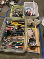 Overview of multiple kitchen utensils in plastic bins and on cutting boards including rolling pin, measuring cups, jar opener, ladle, whisk, flour sifter, and more