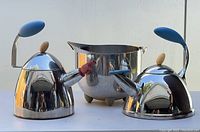 Photo showing two Michael Graves designed stainless steel tea kettles with blue handles, one missing a spinner, and a stainless steel mixing bowl with feet, visible scratches and wear.